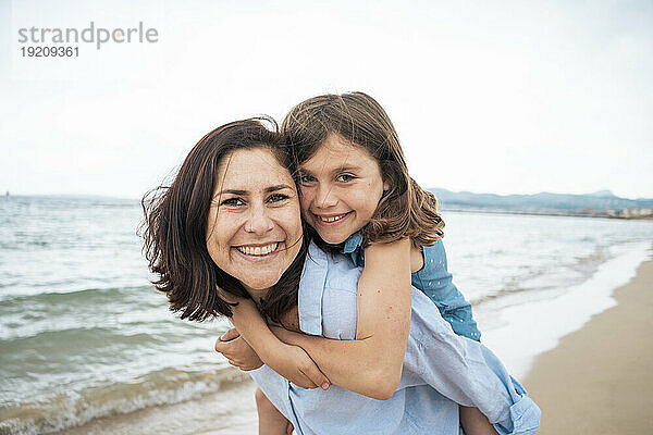 Glückliche Mutter mit Mädchen genießt Huckepackfahrt am Strand