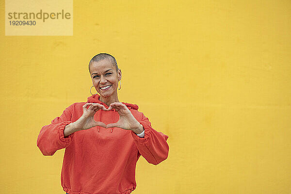 Lächelnde Frau mit rasiertem Kopf  die vor einer gelben Wand ein Herz formt