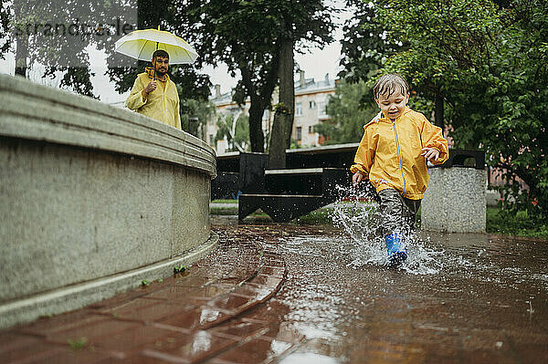 Sohn im Regenmantel spritzt Wasser  Vater im Hintergrund