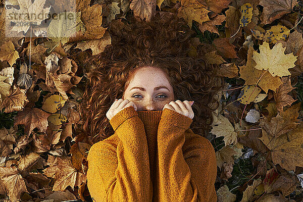 Redhead woman covering face with sweater lying on maple leaves at park