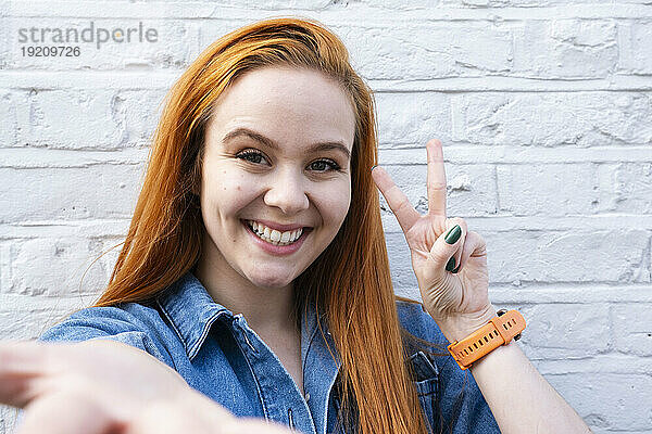 Smiling businesswoman showing peace sign in front of wall