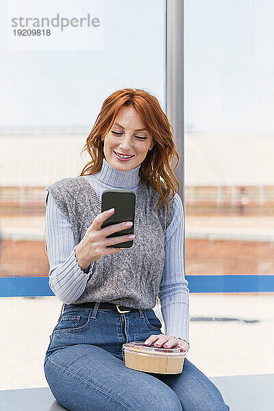 Happy woman using smart phone and sitting with box
