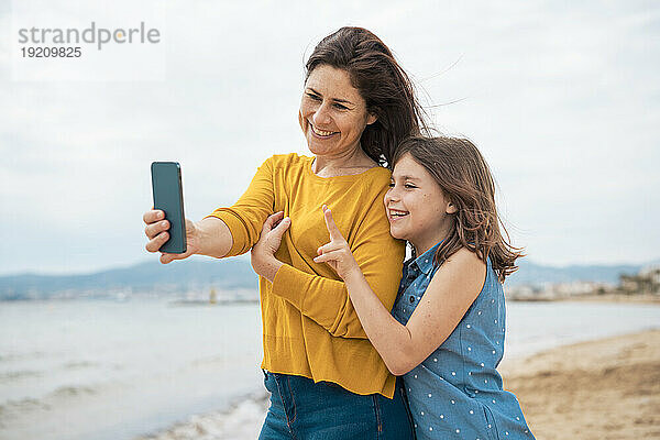 Glückliche Mutter macht ein Selfie mit dem Smartphone  während ihre Tochter am Strand ein Friedenszeichen zeigt