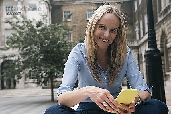 Glückliche Geschäftsfrau sitzt mit Smartphone vor dem Gebäude