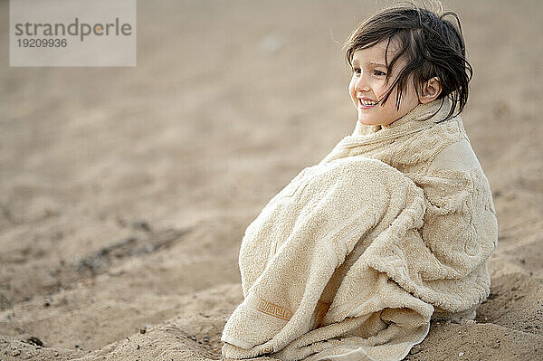 Glücklicher Junge  eingewickelt in ein Handtuch  sitzt auf Sand am Strand