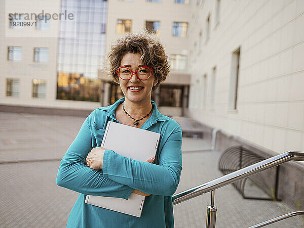 Lächelnde Geschäftsfrau hält Laptop am Geländer gelehnt