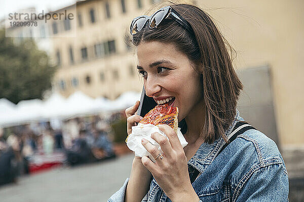 Lächelnde Frau  die Pizza isst und auf dem Smartphone spricht