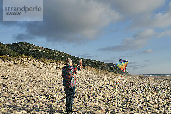 Älterer Mann lässt an einem sonnigen Tag am Strand Drachen unter bewölktem Himmel steigen