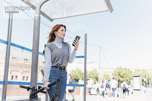 Rothaarige Frau mit Smartphone und Elektroroller an sonnigem Tag