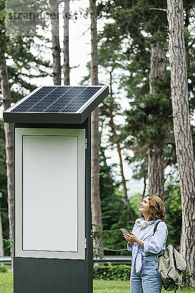 Lächelnde Frau hält Smartphone in der Hand und blickt auf Solarladestation im Park