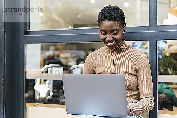 Glückliche junge Frau  die vor dem Café einen Laptop benutzt