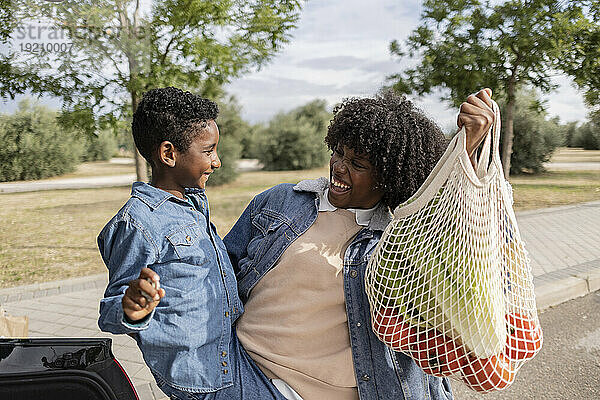 Glücklicher Junge mit Mutter  die an einem sonnigen Tag eine Einkaufstüte trägt