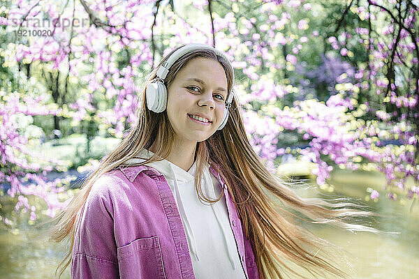 Lächelndes Teenager-Mädchen mit langen blonden Haaren  das Musik mit Kopfhörern am See im Park genießt