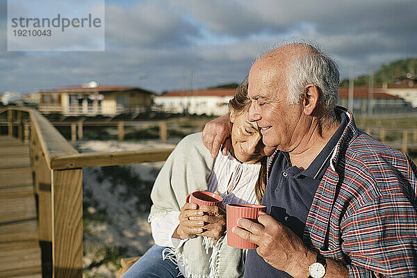 Glückliches älteres Paar hält Kaffeetassen in der Hand und umarmt sich an einem sonnigen Tag