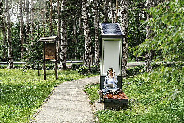 Lächelnde Frau verbringt ihre Freizeit damit  in der Nähe einer Solarladestation im Park zu sitzen