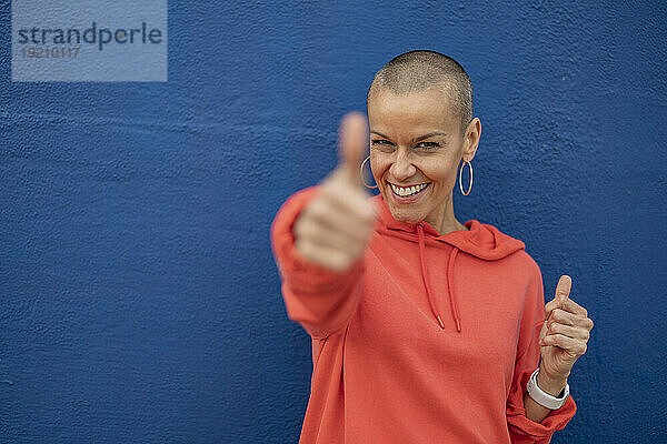 Smiling woman with shaved head showing thumbs up in front of blue wall