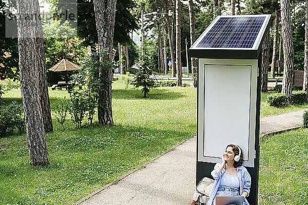 Lächelnde Frau genießt es  Musik zu hören  während sie in der Nähe einer Solarladestation im Park sitzt