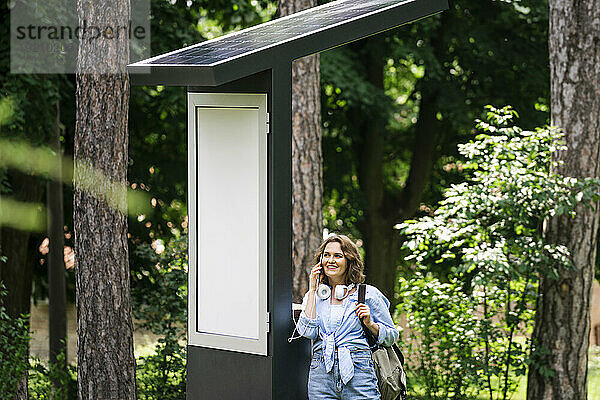 Lächelnde Frau  die mit ihrem Smartphone spricht und in der Nähe einer Solarladestation im Park steht