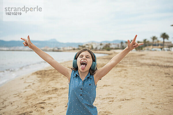 Fröhliches Mädchen mit kabellosen Kopfhörern gestikuliert am Strand mit einem Hupenzeichen