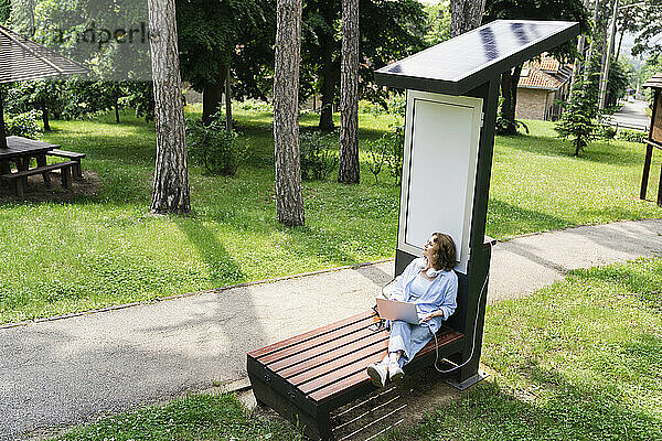 Frau lädt Laptop über Solarladestation im Park auf