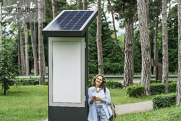 Lächelnde Frau mit Smartphone steht in der Nähe der Solarladestation im Park