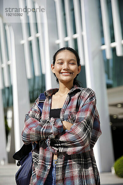 Smiling student with arms crossed standing in front of university