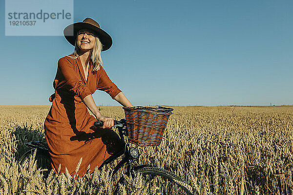 Lächelnde Frau mit Hut fährt Fahrrad im Weizenfeld