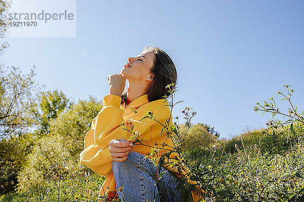Smiling woman with hand on chin sitting in nature under sky