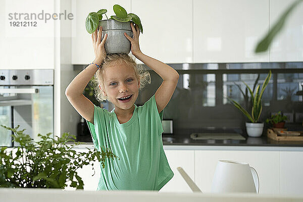 Happy girl balancing potted plant on head in kitchen at home