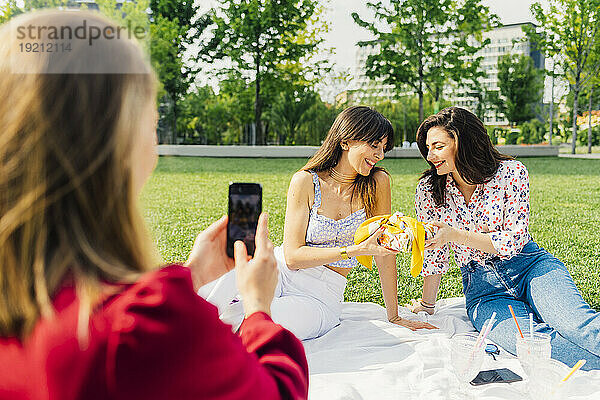 Frau fotografiert Freunde  die auf einer Picknickdecke im Park sitzen