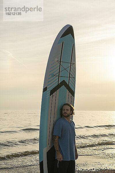 Junger Mann steht vor einem Paddleboard am Strand unter freiem Himmel