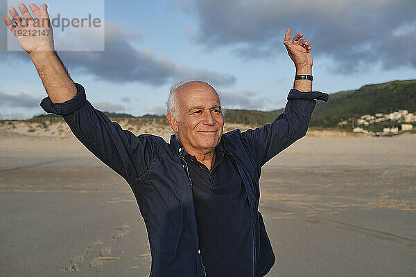 Lächelnder älterer Mann mit erhobenen Armen steht am Strand