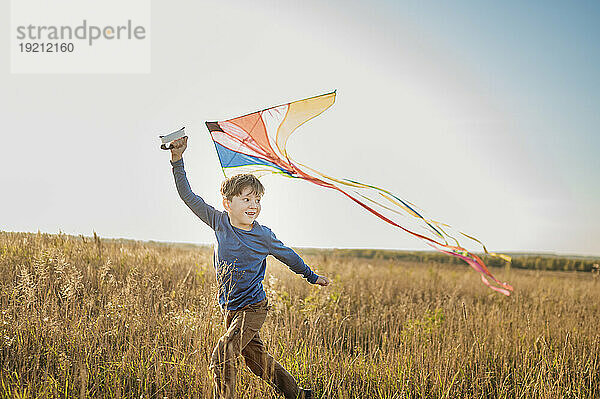 Smiling boy flying kite and running in field under sky