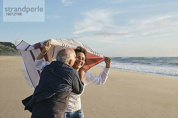 Älterer Mann küsst glückliche Frau mit Schal am Strand