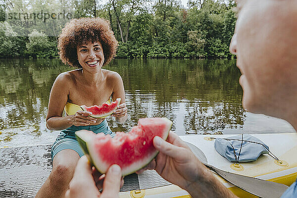 Lächelnde Frau isst Wassermelone mit Freundin auf dem Paddleboard im See