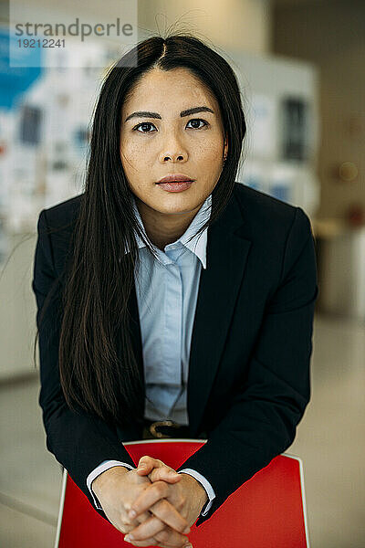 Confident beautiful businesswoman with hands clasped leaning on red chair at office