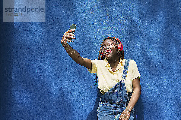 Glückliche junge Frau macht ein Selfie mit ihrem Smartphone vor einer blauen Wand