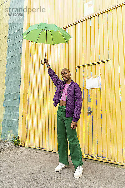 Nicht-binäre Person hält Regenschirm vor gelber Fensterladentür