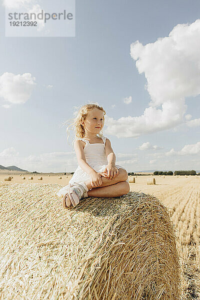 Blondes Mädchen sitzt an einem sonnigen Tag auf einem Strohballen im Feld