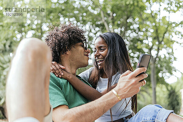 Lächelnde Freundin umarmt Freund mit Smartphone im Park