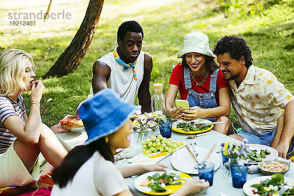 Glückliche Frau zeigt Freunden am Picknicktisch ihr Mobiltelefon
