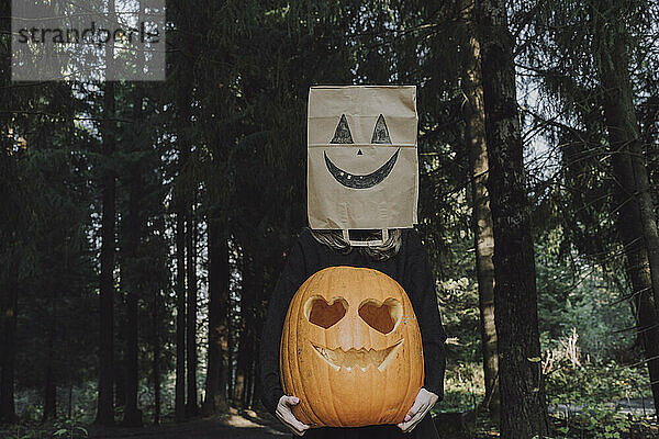 Frau trägt Papiertüte vor Gesicht und hält Jack O'Lantern im Wald