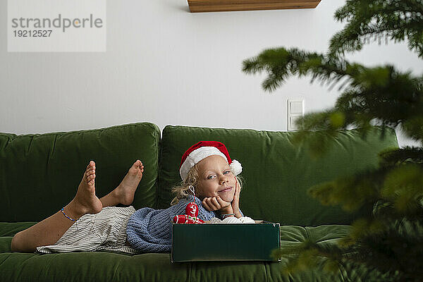 Lächelndes Mädchen liegt auf dem Sofa mit einer Schachtel Weihnachtsschmuck