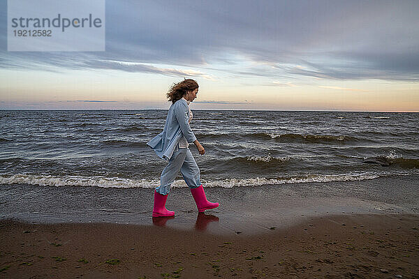 Junge Frau trägt rosa Gummistiefel und geht am Strand spazieren