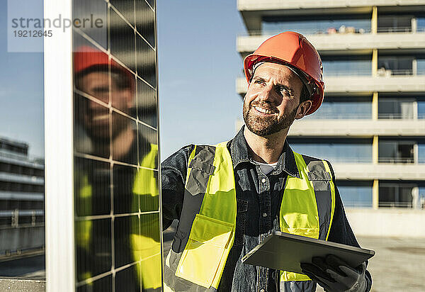 Ingenieur trägt schützende Arbeitskleidung und hält an einem sonnigen Tag ein digitales Tablet in der Hand  das das Solarpanel untersucht