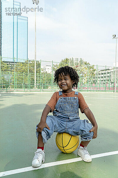 Playful boy sitting on basketball on sports court