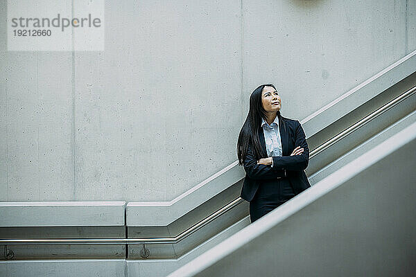 Businesswoman with arms crossed leaning on staircase railing at workplace