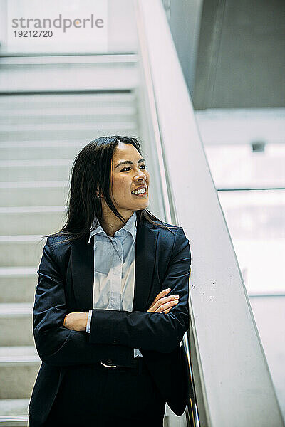 Smiling beautiful businesswoman with arms crossed leaning on staircase railing at office