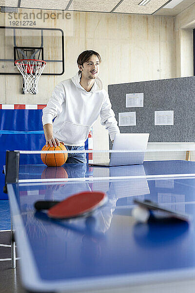 Lächelnder junger Geschäftsmann steht mit Basketball und Laptop am Tennistisch