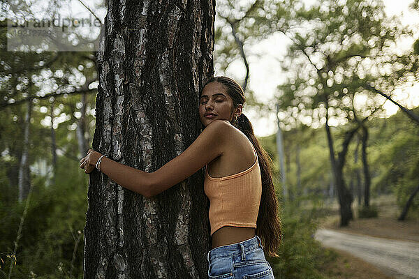 Teenager-Mädchen mit geschlossenen Augen umarmt Baumstamm im Wald bei Sonnenuntergang